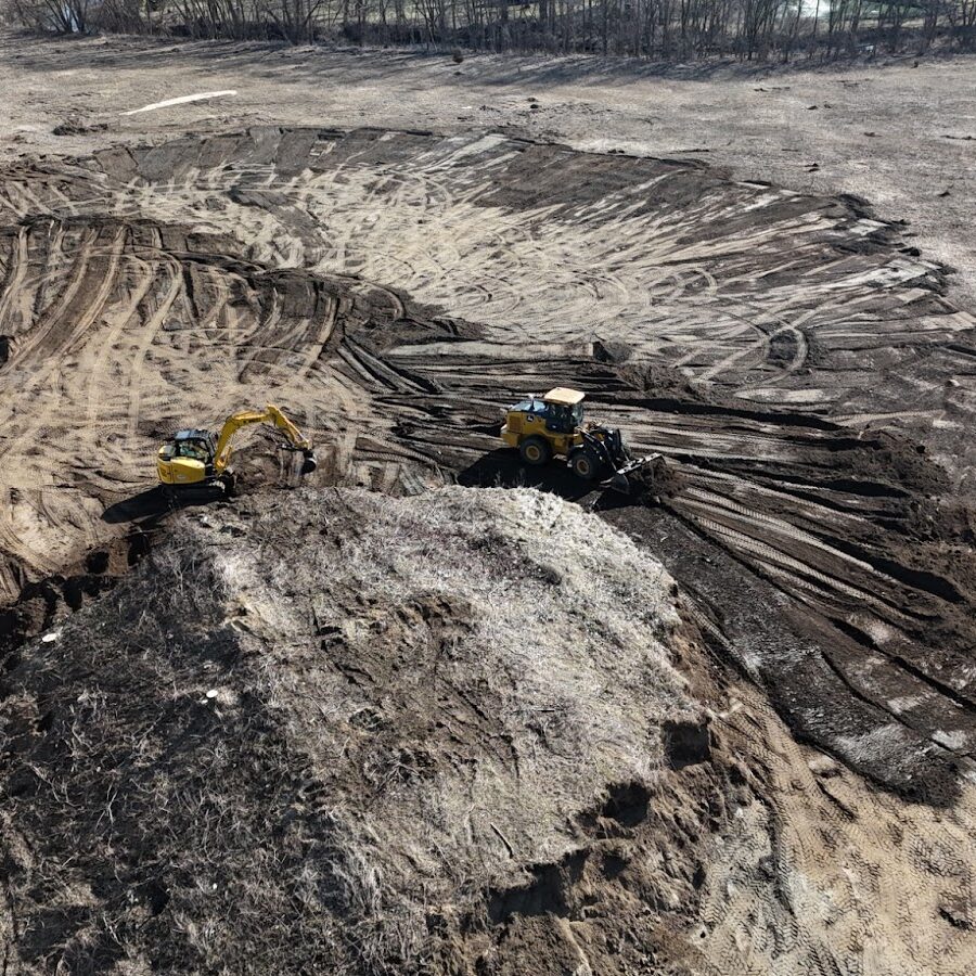 Development Services Aerial view of a construction site with excavators and loaders moving dirt and leveling the ground, leaving visible tire and track marks across the cleared area.