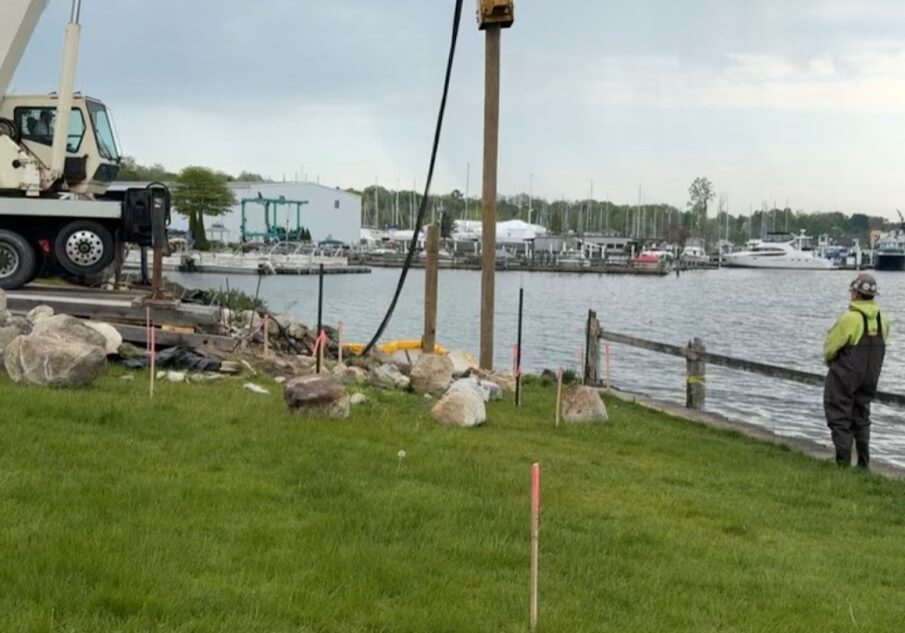 General Contracting Management Crane driving a wooden piling into the ground near a waterfront, with a worker observing and boats docked in the background under a cloudy sky.