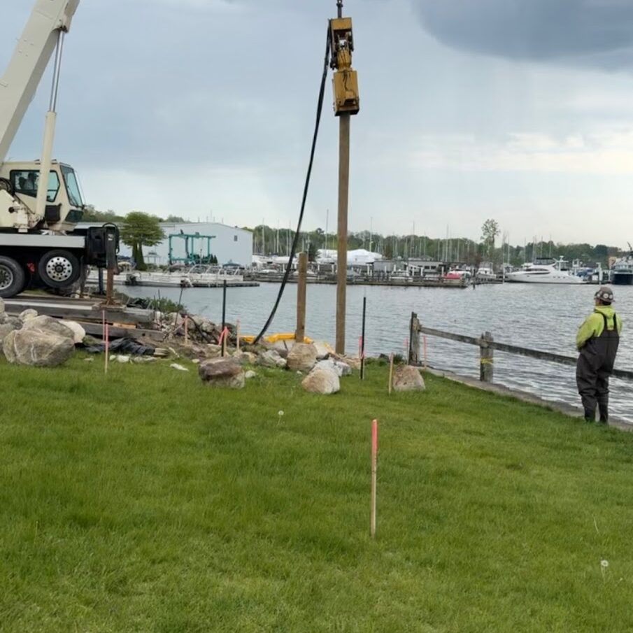 General Contracting Management Crane driving a wooden piling into the ground near a waterfront, with a worker observing and boats docked in the background under a cloudy sky.